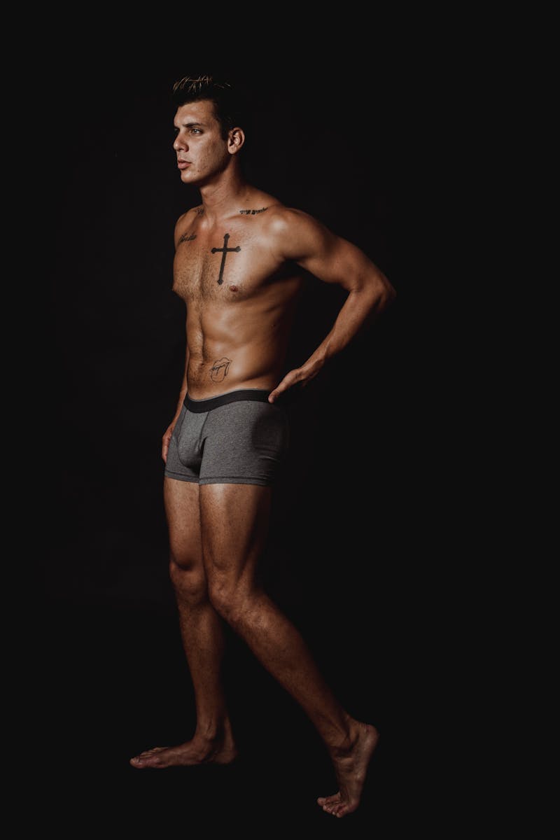 Muscular man with tattoos posing shirtless in a studio setting, emphasizing fitness and strength.